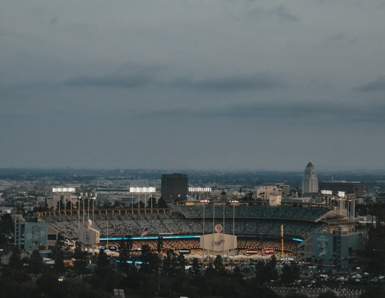 Vista panorámica del estadio de béisbol al anochecer, con las luces encendidas y el horizonte de la ciudad de Los Ángeles de fondo. El estadio está rodeado de edificios y árboles, creando un ambiente urbano y deportivo.