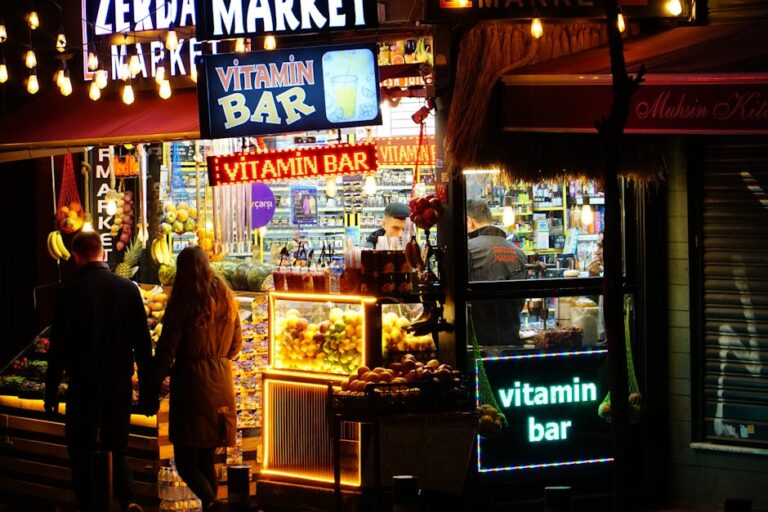Imagen de un mercado iluminado por luces nocturnas, con un letrero que dice "Vitamin Bar". Se ven frutas frescas en la ventana, y dos personas de espaldas observan el lugar. El ambiente es vibrante y acog