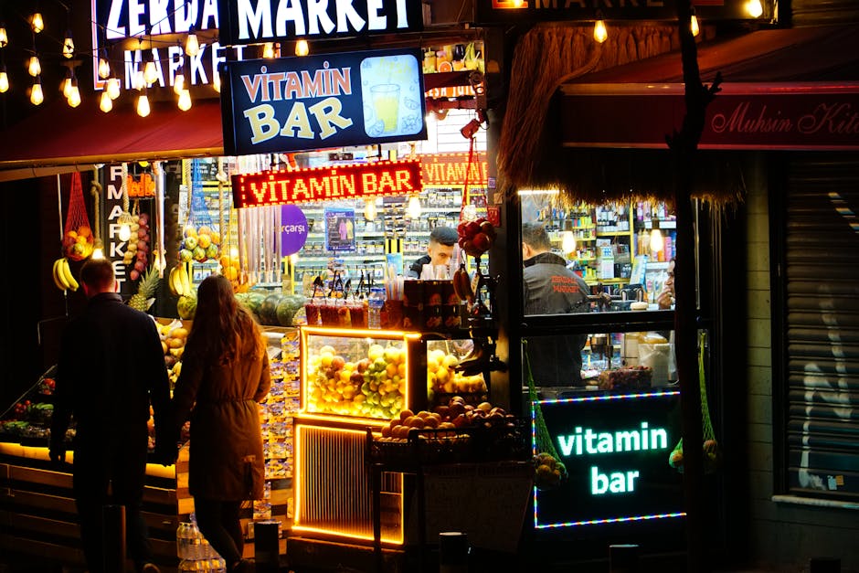Imagen de un mercado nocturno con un letrero brillante que dice "Vitamin Bar". La escena muestra a dos personas de espaldas, frente a un puesto lleno de frutas frescas y luces coloridas. Al fondo, se puede ver a los