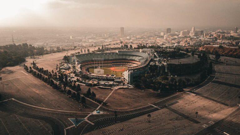Vista aérea del Dodger Stadium en Los Ángeles, rodeado de un paisaje urbano y un vasto estacionamiento. El cielo presenta un tono nublado, creando una atmósfera dramática sobre el estadio.