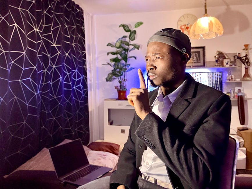 Hombre con traje negro y gorra, sentado en un ambiente interior moderno, pidiendo silencio con un dedo en los labios. Fondo decorado con plantas y una lámpara suave. Un ordenador portátil cerrado en la esquina.