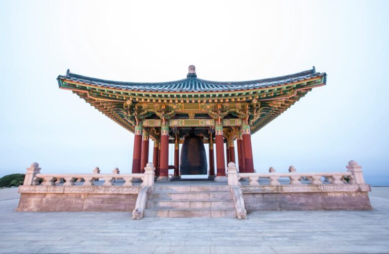 Templo tradicional asiático con tejado curvo y decoraciones coloridas, sosteniendo una gran campana en el centro. La estructura está rodeada de barandas y se encuentra en un entorno sereno, ideal para actividades culturales y ceremoniales