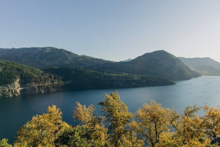 Paisaje sereno que muestra un lago rodeado de montañas y árboles con hojas amarillas, bajo un cielo despejado. Ideal para transmitir tranquilidad y belleza natural.