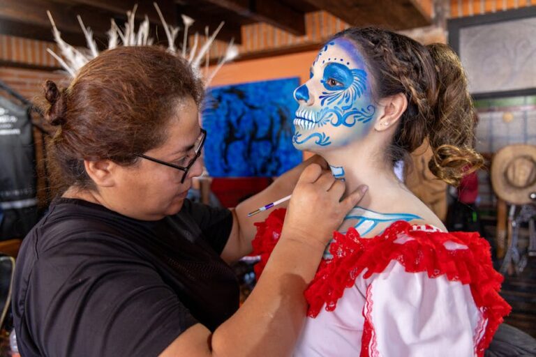 Retrato de una mujer recibiendo un maquillaje artístico de calavera, típico del Día de Muertos, mientras otra persona aplica detalles en su rostro. La modelo lleva un vestido tradicional mexicano con detalles rojos, y el fondo muestra decoraciones color