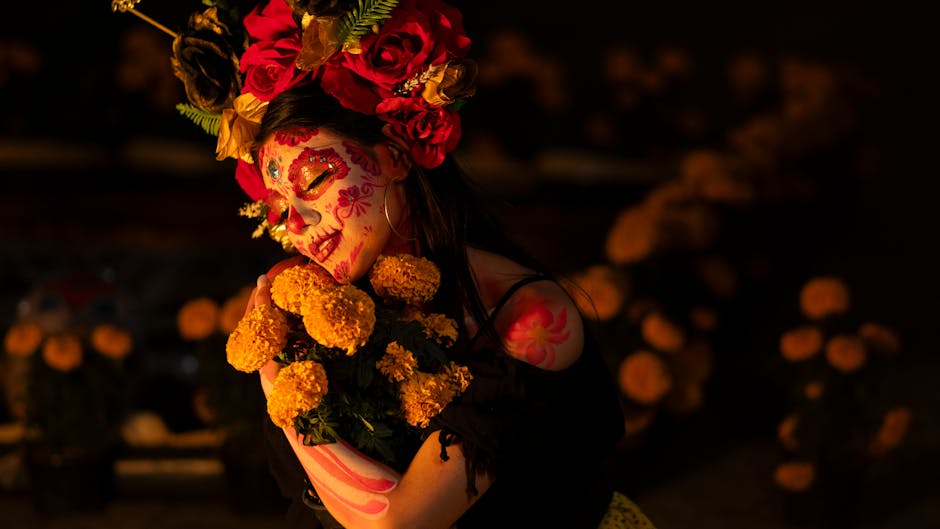 Retrato de una mujer con maquillaje tradicional de calavera, sosteniendo un ramo de flores amarillas. Luce un tocado de flores brillantes y un fondo oscuro con decoraciones de flores de cempasúchil, evocando la celebración