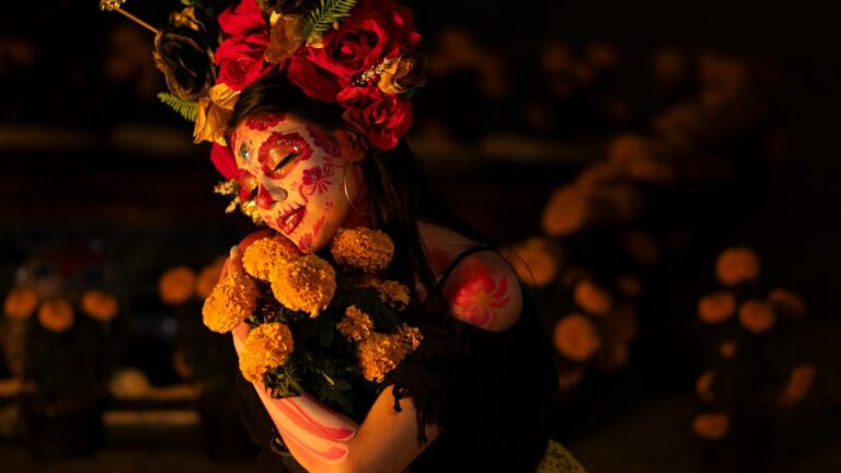 Mujer con maquillaje de calavera y un tocado floral, sosteniendo un ramo de flores amarillas. La imagen es un homenaje visual a la celebración del Día de Muertos, con un fondo oscuro y decoraciones florales.