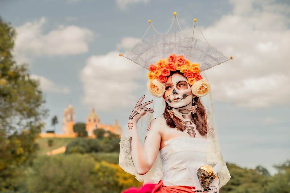 Mujer con maquillaje de calavera, vestida con un atuendo blanco y rojo, posa con una mano en alto. Lleva una corona de flores de color naranja y un velo, mientras sostiene un cráneo dorado. Al fondo,