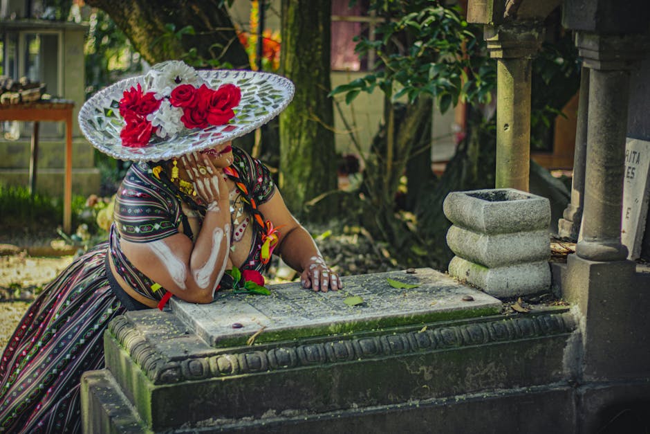 Mujer con un sombrero decorado con flores rojas, posando pensativa sobre una lápida en un cementerio. Viste una blusa tradicional y presenta pintura blanca en los brazos, rodeada de naturaleza y monumentos funerarios