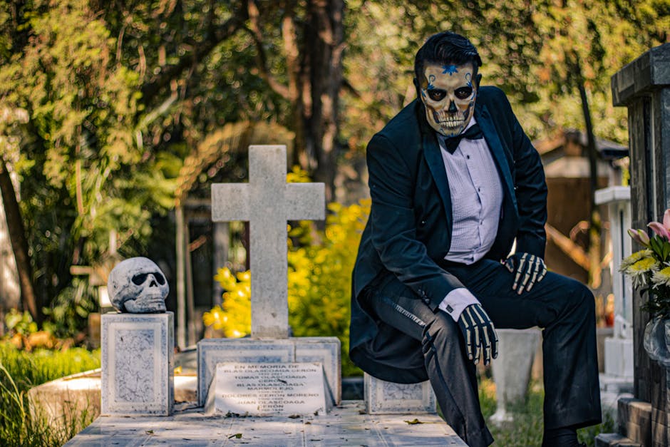 Hombre con maquillaje de calavera sentado en una lápida en un cementerio, vestido de traje negro y corbata, con un fondo de naturaleza y elementos funerarios como cruz y calavera. La imagen evoca la celebración del Día de