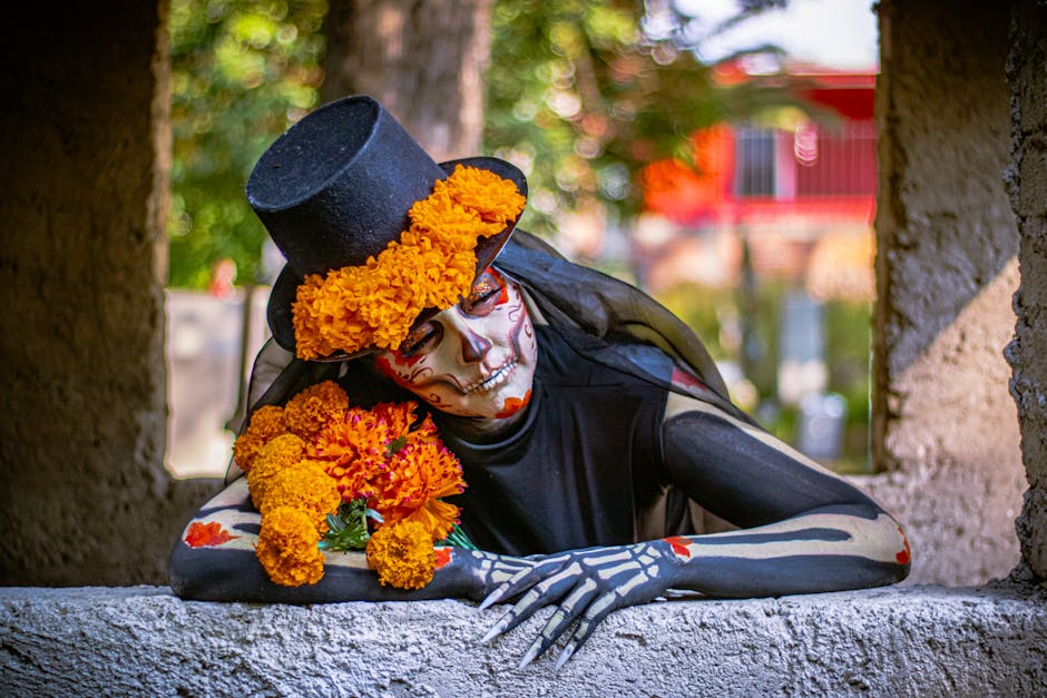 Persona vestida de calavera con maquillaje detallado, sombrero de copa y flores de cempasúchil, posando sobre un muro en un ambiente natural, representando el Día de Muertos.