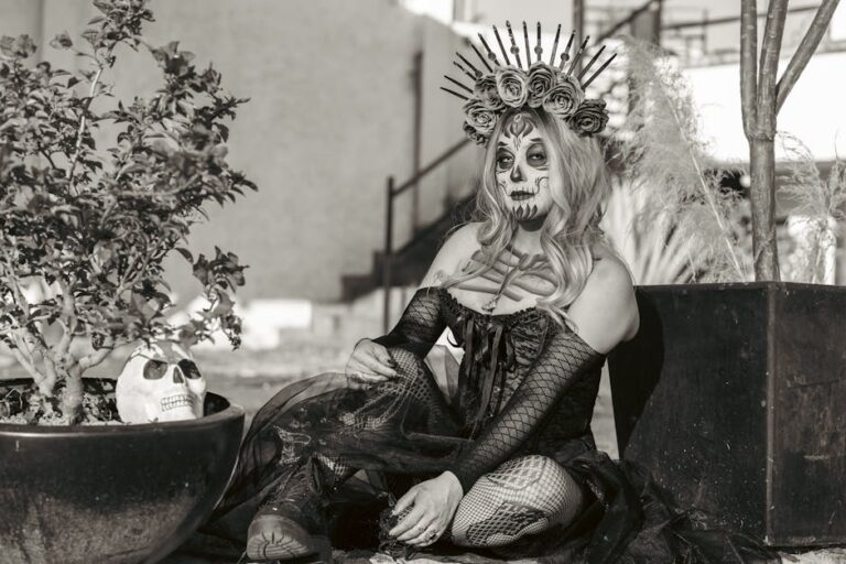 Mujer con maquillaje de calavera y vestido oscuro, sentada junto a plantas en macetas. Lleva una corona de flores y una decoración de espinas. Imagen en blanco y negro que evoca un estilo de Día de Muertos.