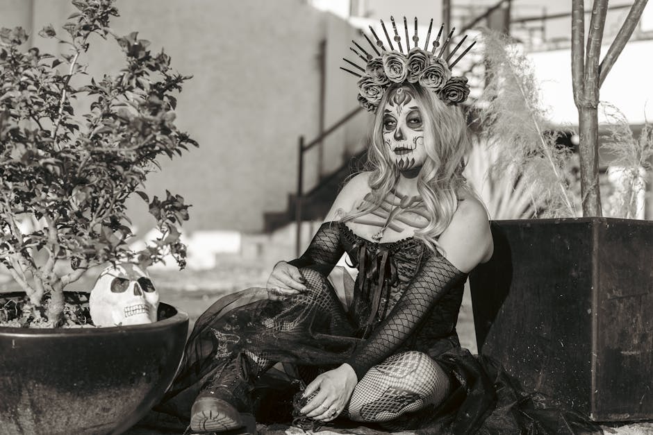 Mujer con maquillaje de calavera y vestido oscuro, sentada junto a plantas en macetas. Lleva una corona de flores y una decoración de espinas. Imagen en blanco y negro que evoca un estilo de Día de Muertos.