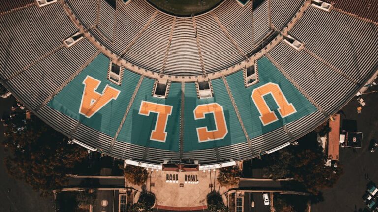 Imagen aérea de un estadio con asientos vacíos y el campo pintado con las letras "UCLA". El área está iluminada por la luz del día, mostrando una vista clara del diseño del campo y la estructura del estadio.
